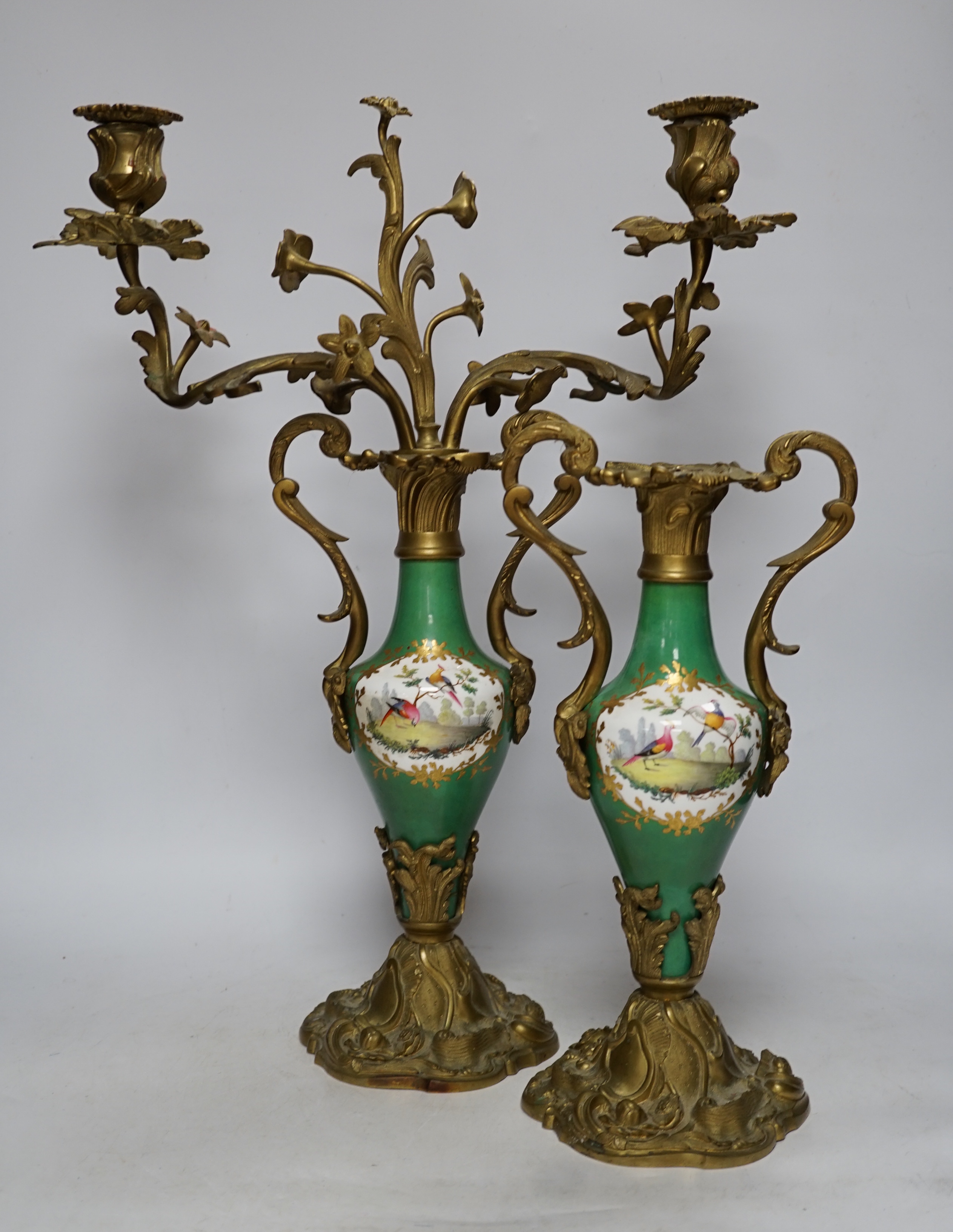 A pair of Sevres style porcelain and brass rococo-style two branch candelabra, hand painted with exotic birds, 50cm high